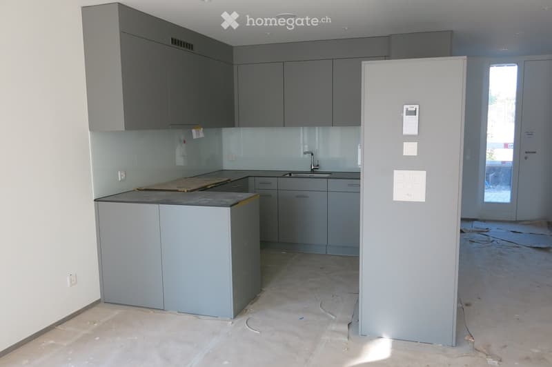 Empty kitchen with grey cabinets, a central island, and a sink area. The floor is unfinished concrete, and the space is well-lit with natural light from a large window.