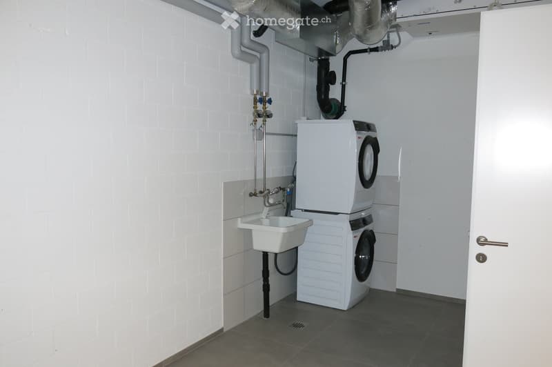 A small, clean utility room with white brick walls, a washing machine, a sink, and a faucet.
