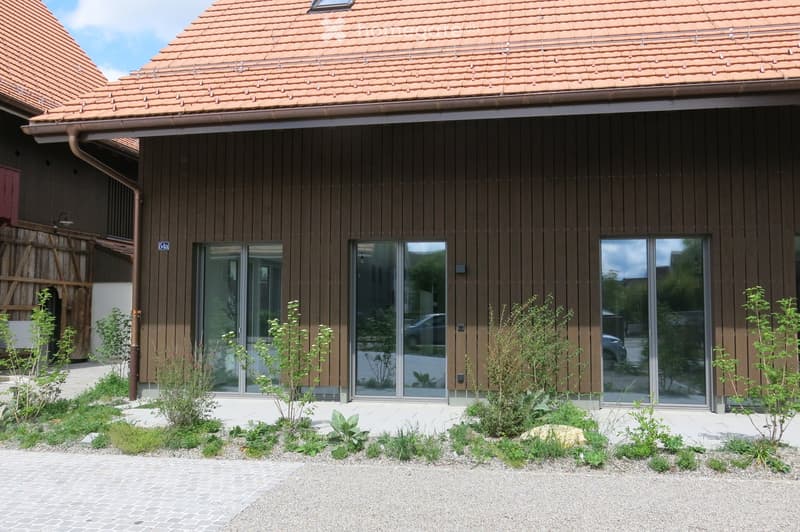 Modern two-story house with brown wooden facade, large glass windows, and a tiled roof. The front yard features small plants and shrubs arranged in a landscaped bed.