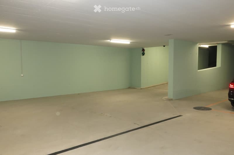 Empty underground parking space with concrete floor, light green walls, fluorescent lighting, and a single car partially visible at the edge.