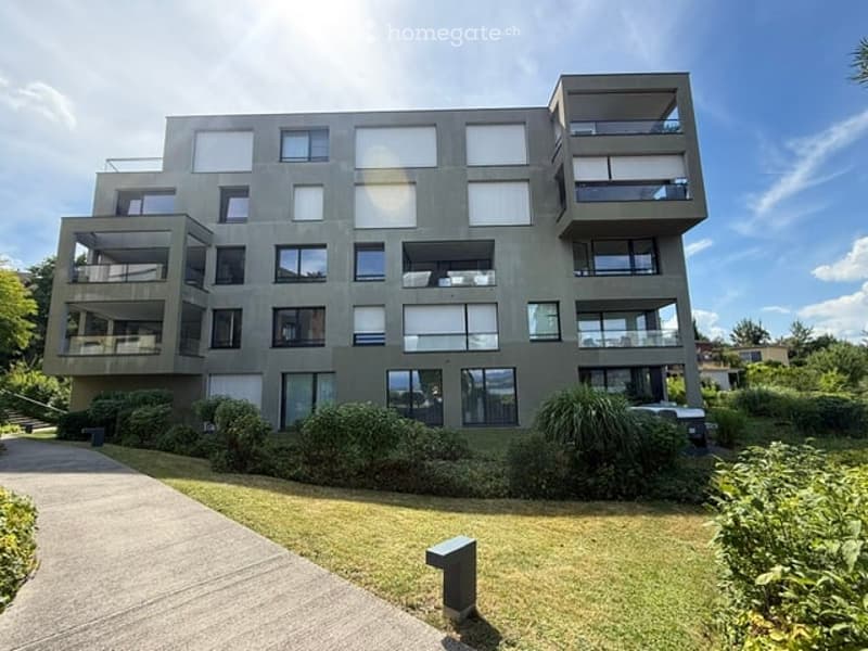 Modern multi-story apartment building with large windows and balconies, surrounded by greenery and a paved walkway.