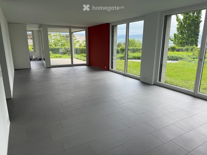 Empty room with grey tiled floor, large windows, and sliding glass doors leading to a garden area.
