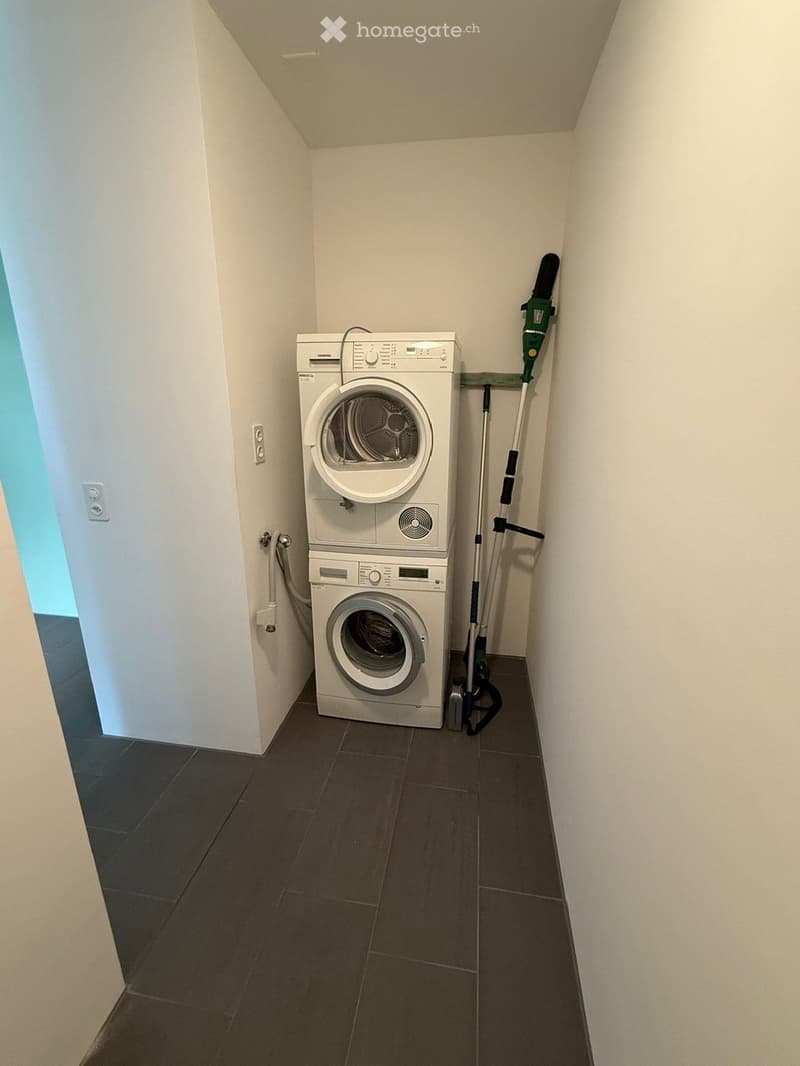 A compact bathroom with a stacked washing machine and dryer unit, dark tiled floor, white walls, and a vacuum cleaner leaning against the wall.