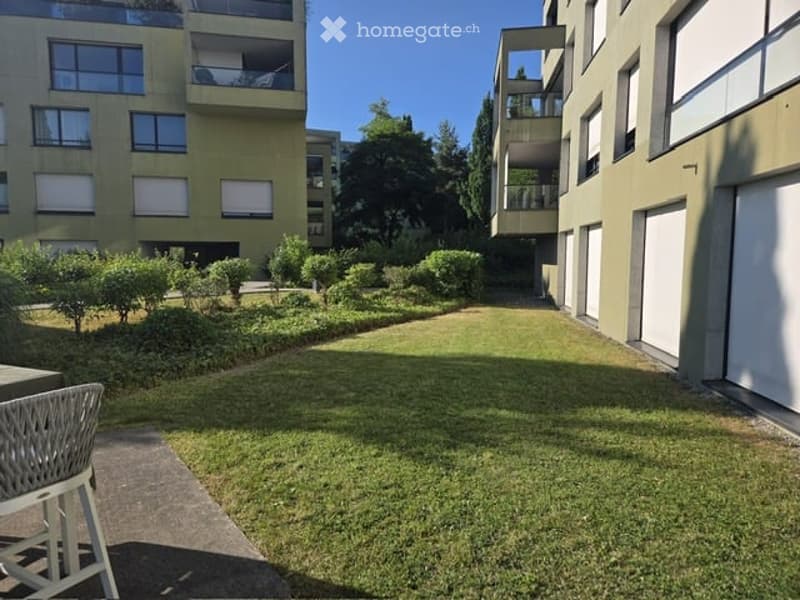 Well-maintained garden area with trimmed bushes, a small lawn, and a few modern apartment buildings in the background. A white wicker chair is visible in the foreground.