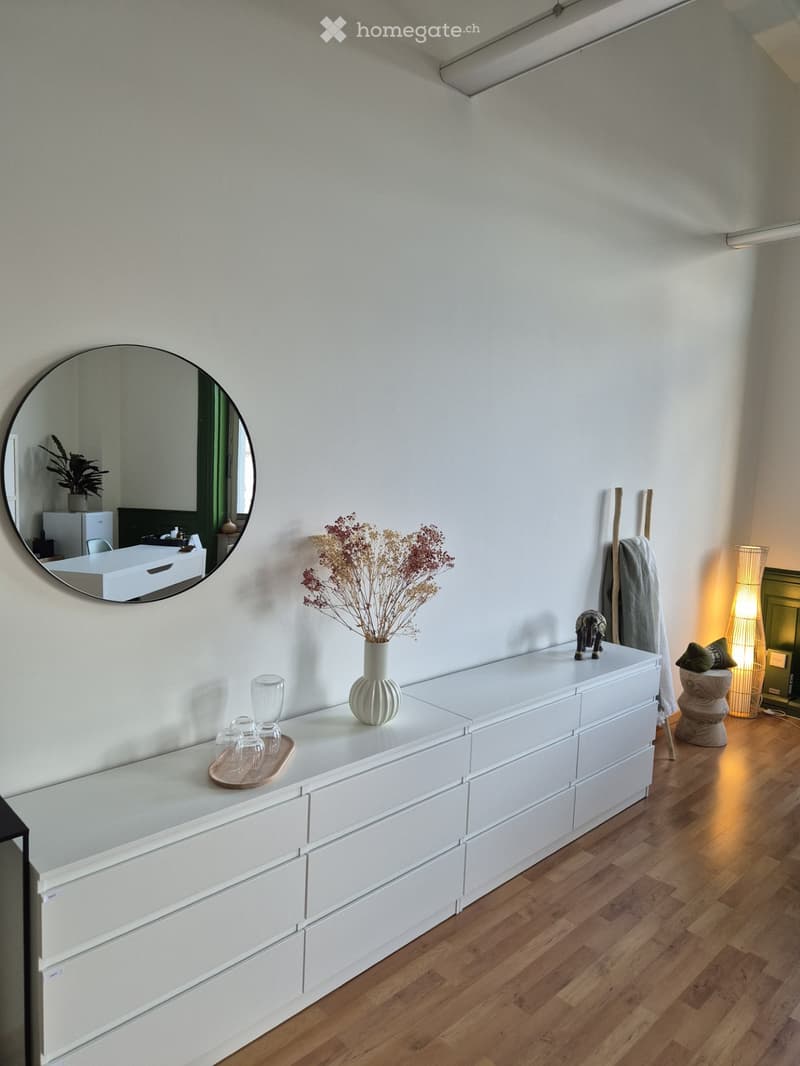 A modern, minimalist room with a white dresser, a vase with dried flowers, a round mirror, and a small table with glassware. The room features light-colored walls and wooden flooring.