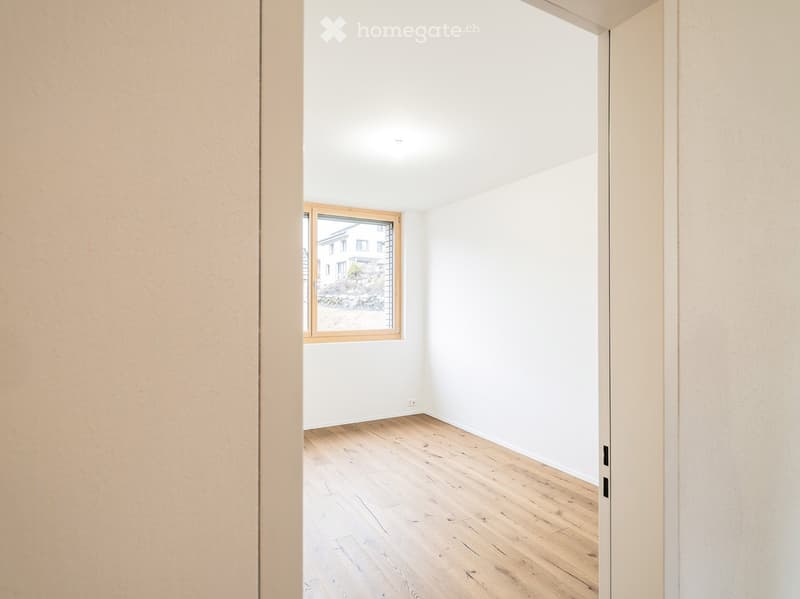 Empty room with light wooden flooring, white walls, and a large window with wooden frame showing a view of the outside.