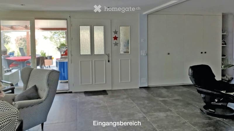 The image shows a modern, well-lit living space with a grey tiled floor, a grey checkered armchair, a black leather chair, and a large white cabinet. There is a large glass door leading to an outdoor area with a red table and chairs.