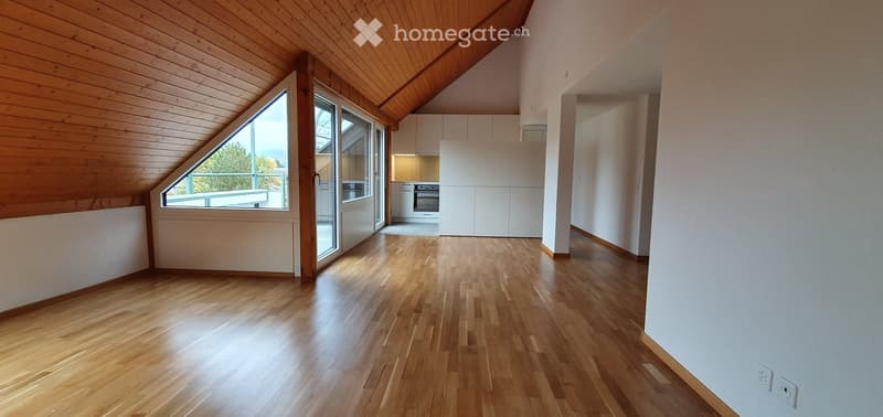 Bright, empty attic space with wooden floors and slanted wooden ceiling. Features large windows and a small, modern kitchen area with white cabinets and stainless steel appliances.