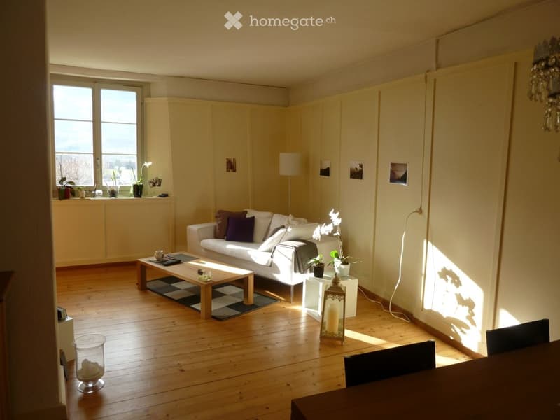 Bright living room with wooden flooring, white walls, large windows, a white sofa with colorful cushions, a coffee table, a dining table with chairs, and decorative items including candles and vases.