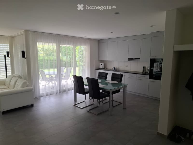 Modern open-plan living and dining area with a white sofa, glass dining table with chairs, and a fitted kitchen with cabinets and appliances.