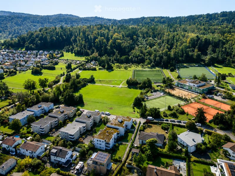 Aerial view of a residential area with multiple houses, green spaces, sports fields, and a forested background.