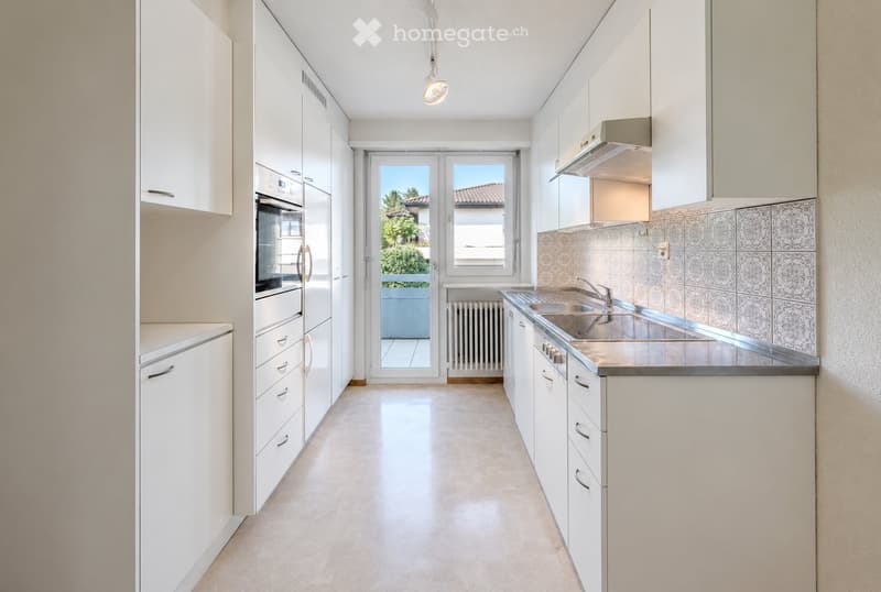 Modern kitchen with white cabinets, stainless steel sink, built-in oven, and a view of a balcony through a glass door.