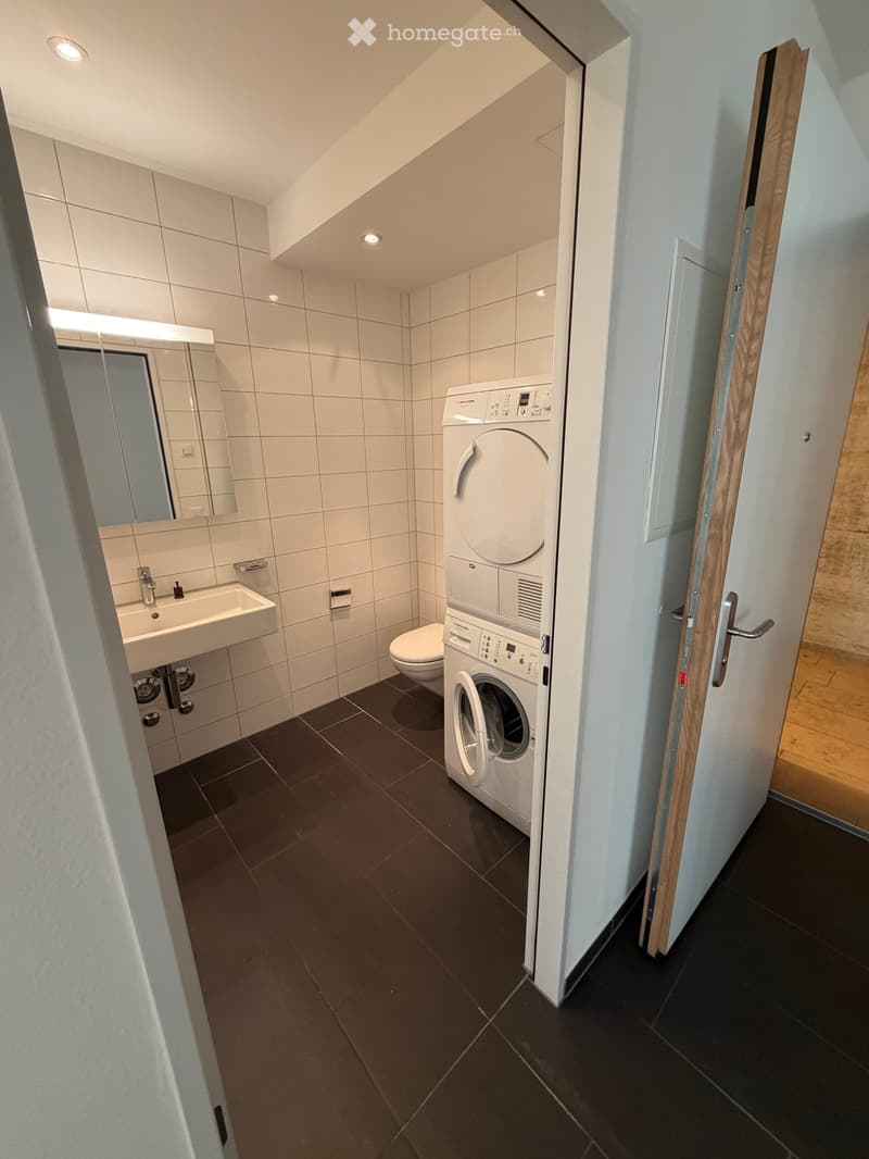 Modern bathroom with white tiles, a sink, mirror, toilet, and a stacked washing machine/dryer unit.