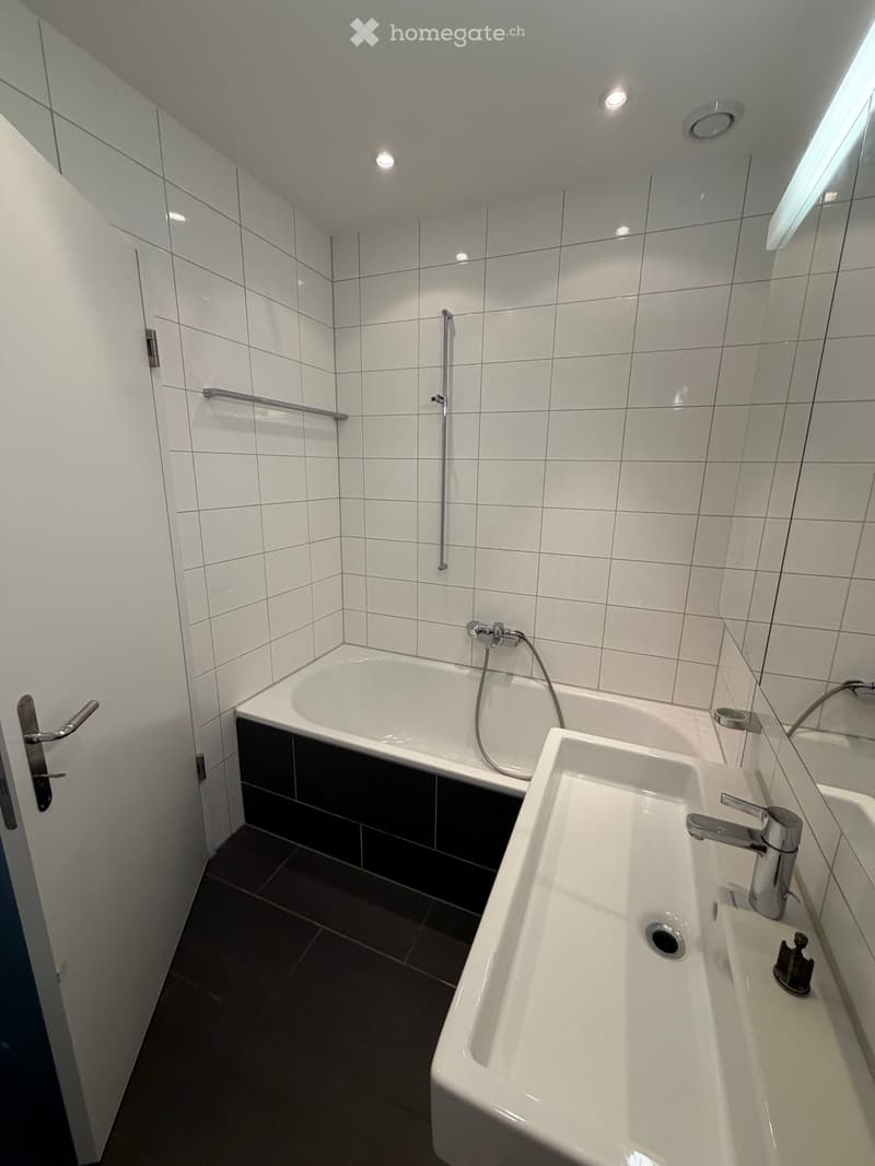 Modern bathroom with white tiled walls, a bathtub, a double sink, and a large mirror. Features include a handheld showerhead, a door with a silver handle, and recessed ceiling lights.