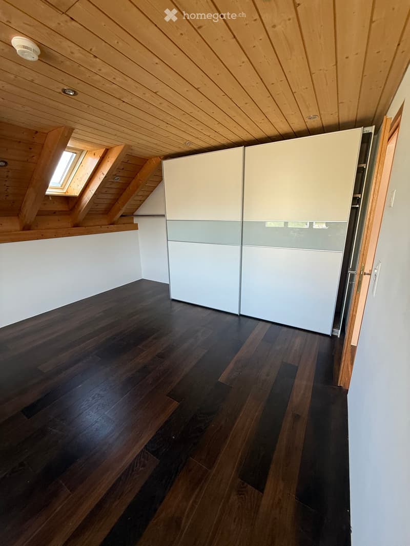 An empty attic room with wooden floors, white walls, and a built-in closet with white and grey doors. The ceiling has exposed wooden beams and a skylight window.