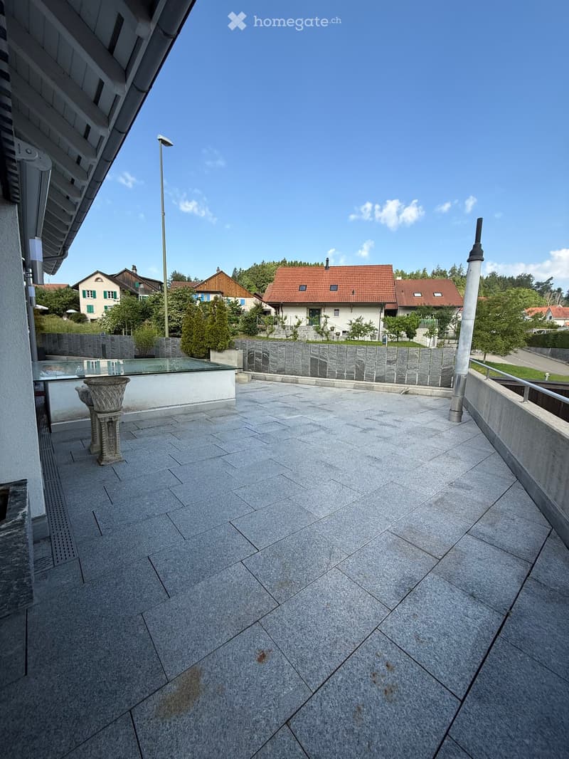 Spacious balcony with grey stone tiles, glass railing, and a decorative urn on the left side. The background shows a residential area with houses and greenery.