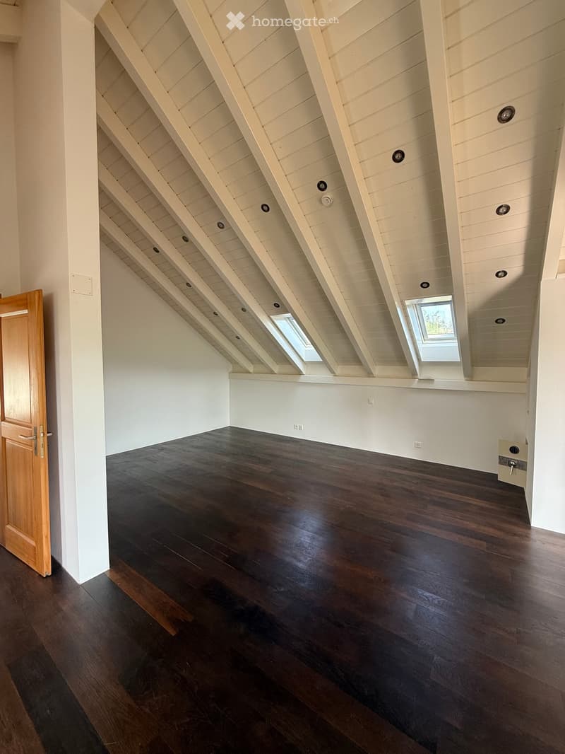 An empty attic space with exposed wooden beams, dark hardwood flooring, and skylight windows, featuring white walls and a wooden door.