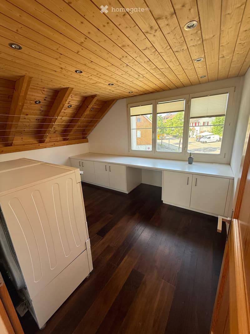Bright attic room with wooden slanted ceiling, dark wooden floor, white cabinets, and a small desk with a potted plant on top.