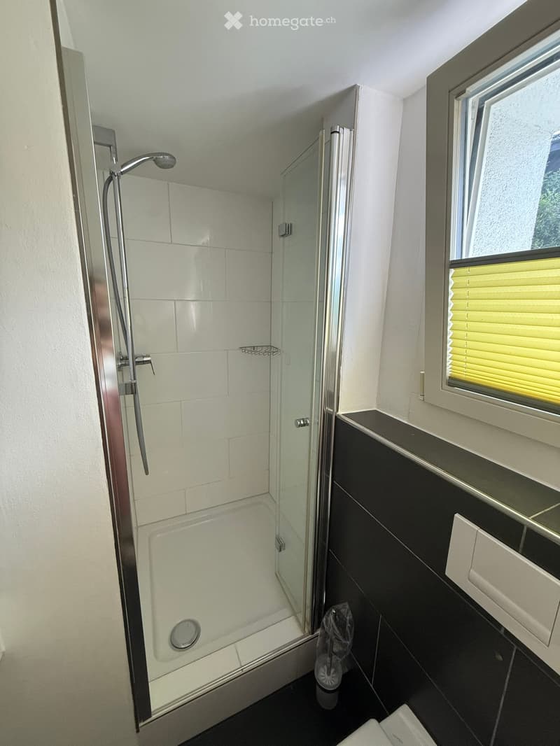 Modern bathroom with a glass-enclosed shower, white tiles, black lower cabinets, and a window with blinds.