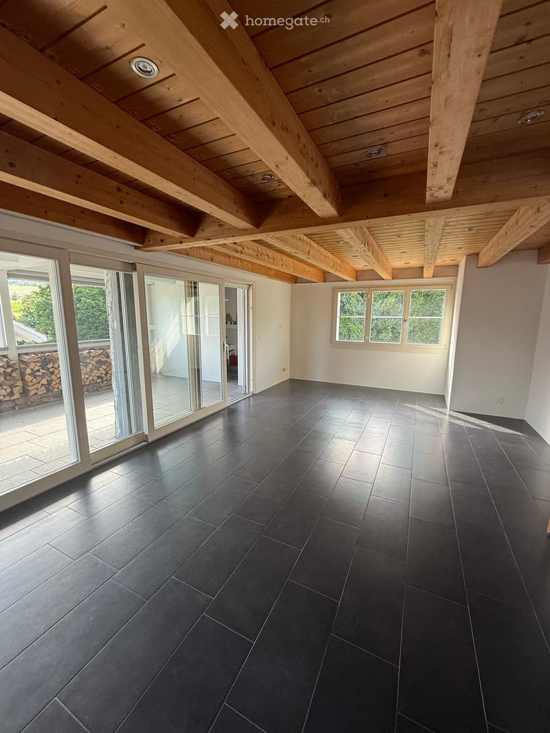 Bright, open room with dark grey tiled floor, exposed wooden ceiling beams, large windows, and sliding glass doors leading to an outdoor area.