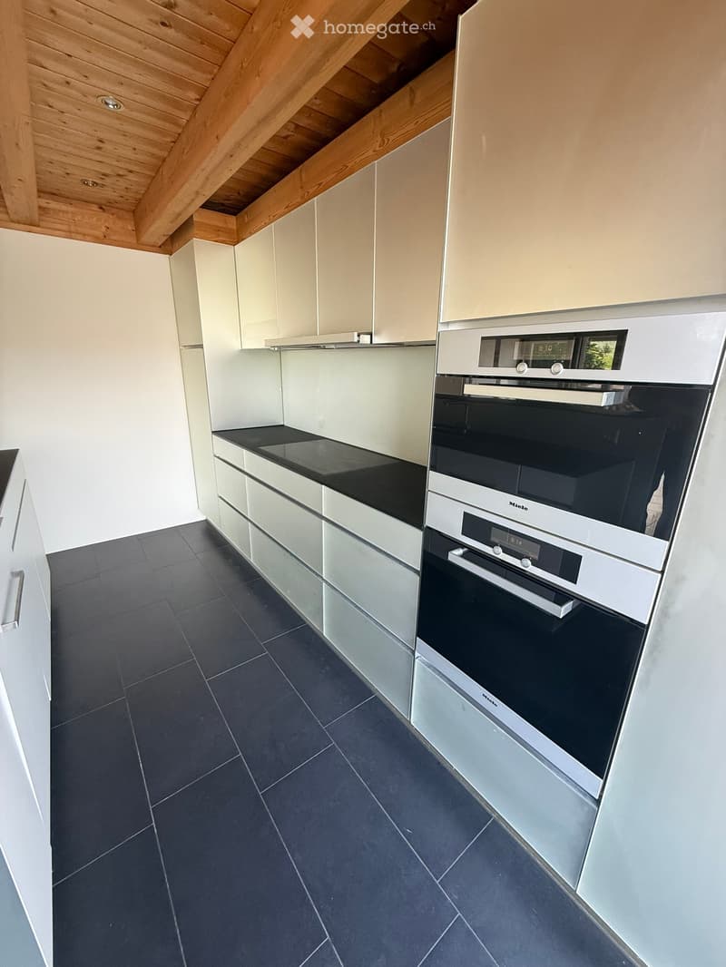 Modern kitchen with white cabinets, black countertop, and a built-in oven. The floor is dark tiled, and the ceiling has exposed wooden beams.