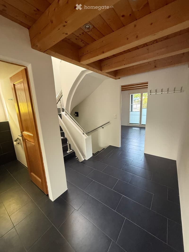 Slanted ceiling hallway with dark tiled floor, white walls, wooden door, staircase with white railing, and an open doorway leading to another room with large windows.