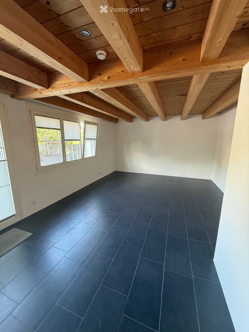 An empty room with exposed wooden beams on the ceiling, dark blue tiles on the floor, and large windows with blinds.