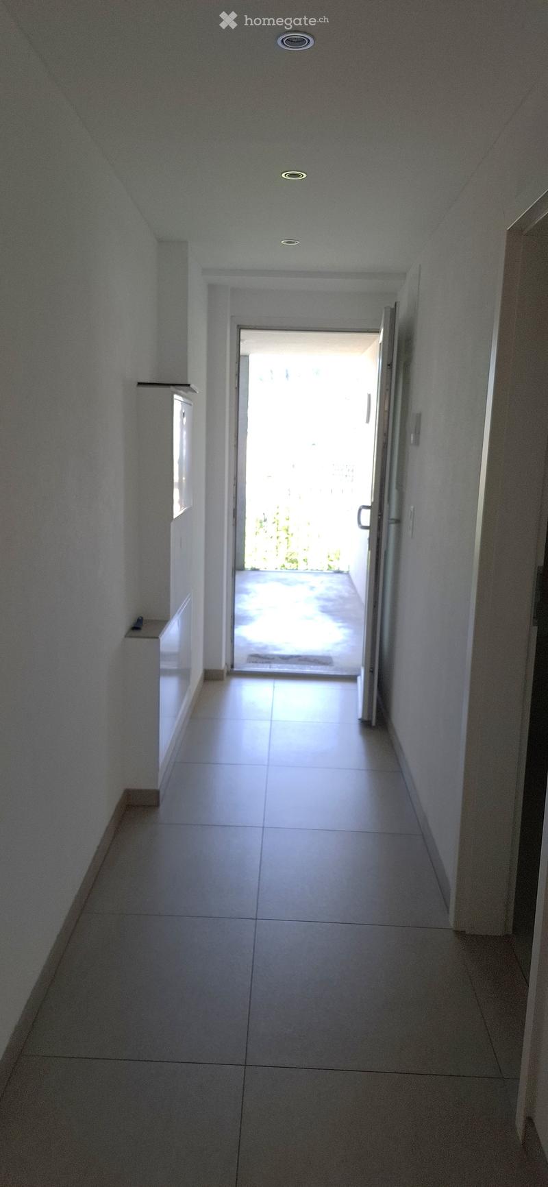 Empty hallway with white tiled flooring, white walls, and recessed ceiling lights. An open door at the end leads to a bright outdoor area with a pool visible.