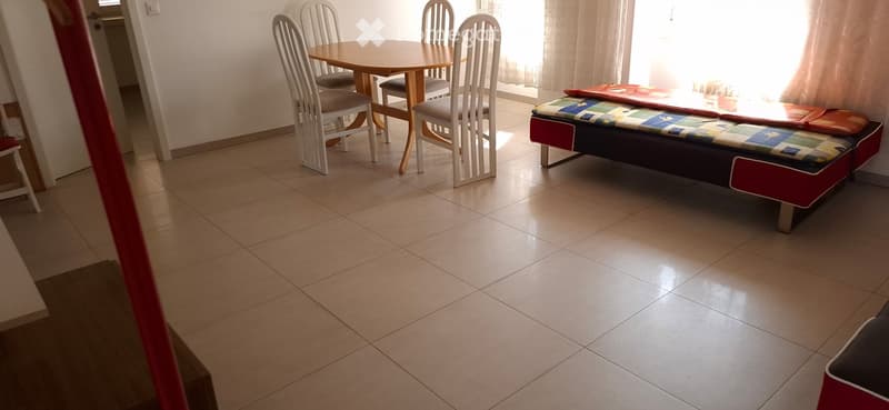 A furnished room with a sofa, a dining table, and chairs, featuring tiled flooring and natural light from a window.