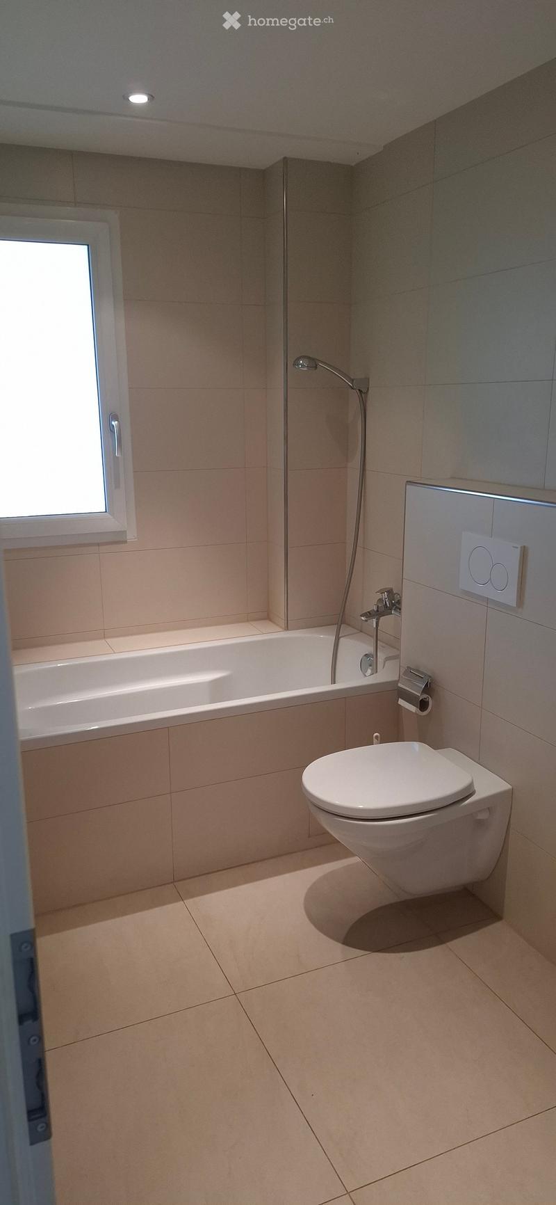Modern bathroom with a bathtub, toilet, and showerhead, featuring beige tiles and a window for natural light.