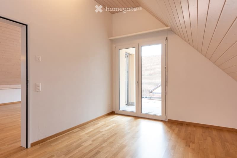 Bright, empty room with wooden flooring and white walls, featuring a large sliding glass door leading to a balcony or outdoor area. The ceiling has a sloped, wooden design typical of an attic space.