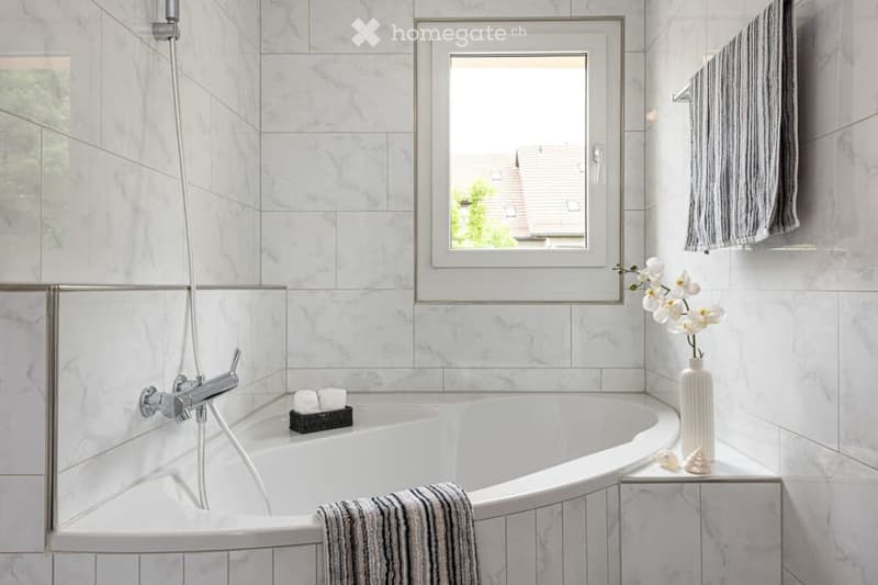 Modern bathroom with marble tiles, a large bathtub, a showerhead, a window, and decorative items including a vase with white flowers and towels.