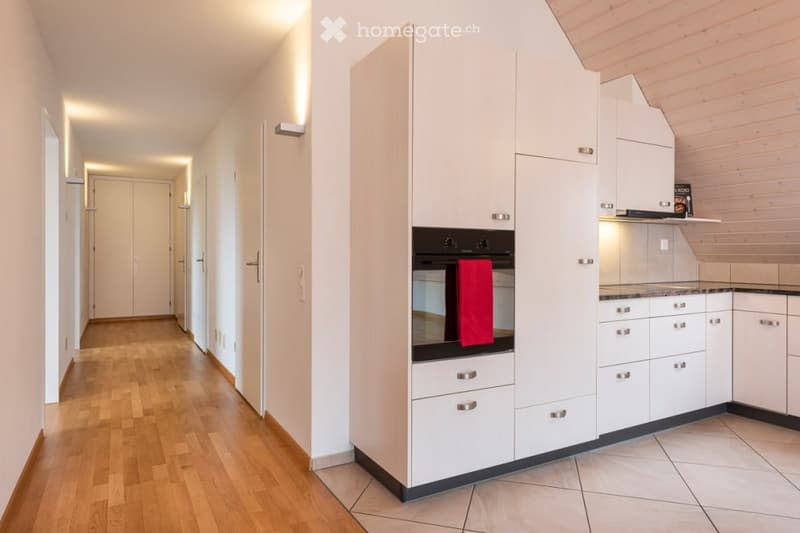 Bright hallway with wooden flooring, white walls, and recessed lighting, leading to a modern kitchen area with white cabinets, stainless steel appliances, and a tiled floor.