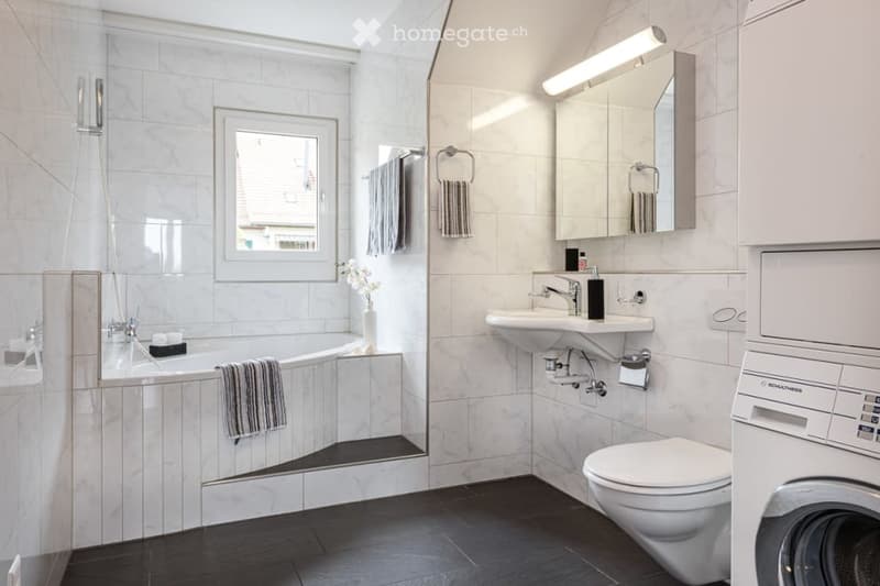 Modern bathroom with white tiles, a bathtub, sink, toilet, mirror, and a washing machine. Features include a window, towels, and a small vase with flowers.