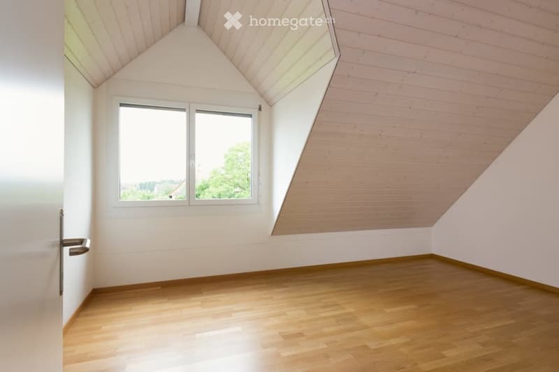 Empty room with wooden flooring, white walls, and a large window offering a view of greenery outside. The ceiling has a sloped, wooden design.