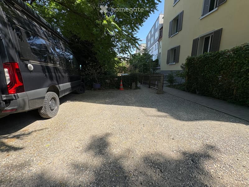 Gravel driveway with a parked van, a small garden area with plants, and a building with multiple windows and shutters in the background.