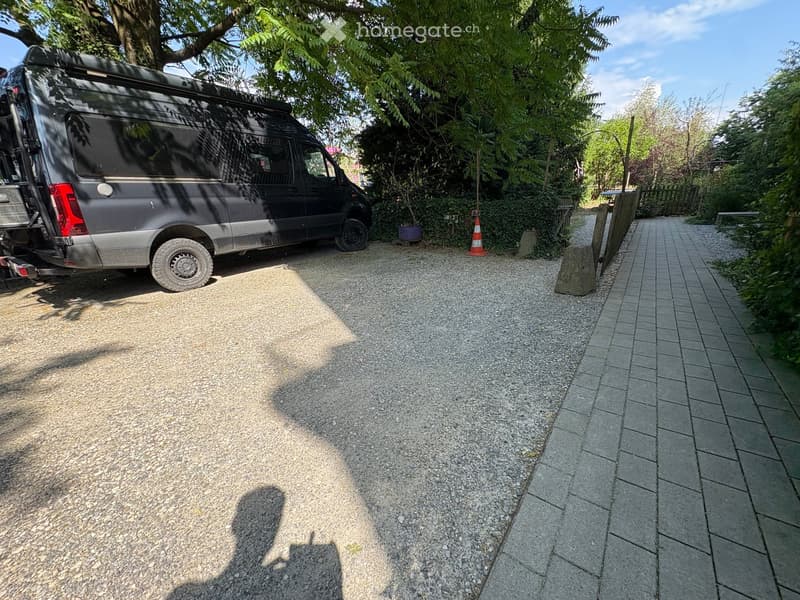 Gravel driveway with a parked van, surrounded by greenery and trees. A paved pathway leads to a wooden fence.