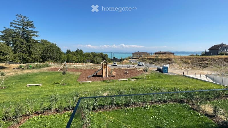 A grassy outdoor area with a small playground featuring a wooden play structure, slide, and benches. The area is bordered by a fence and includes trees and a view of the water in the background.