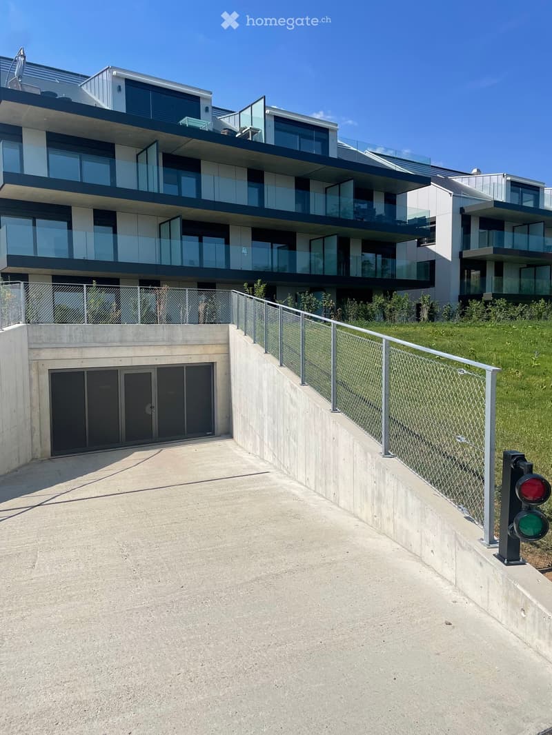 Modern multi-story building with large glass windows and balconies, raised ground floor with concrete walls and a fenced pathway.