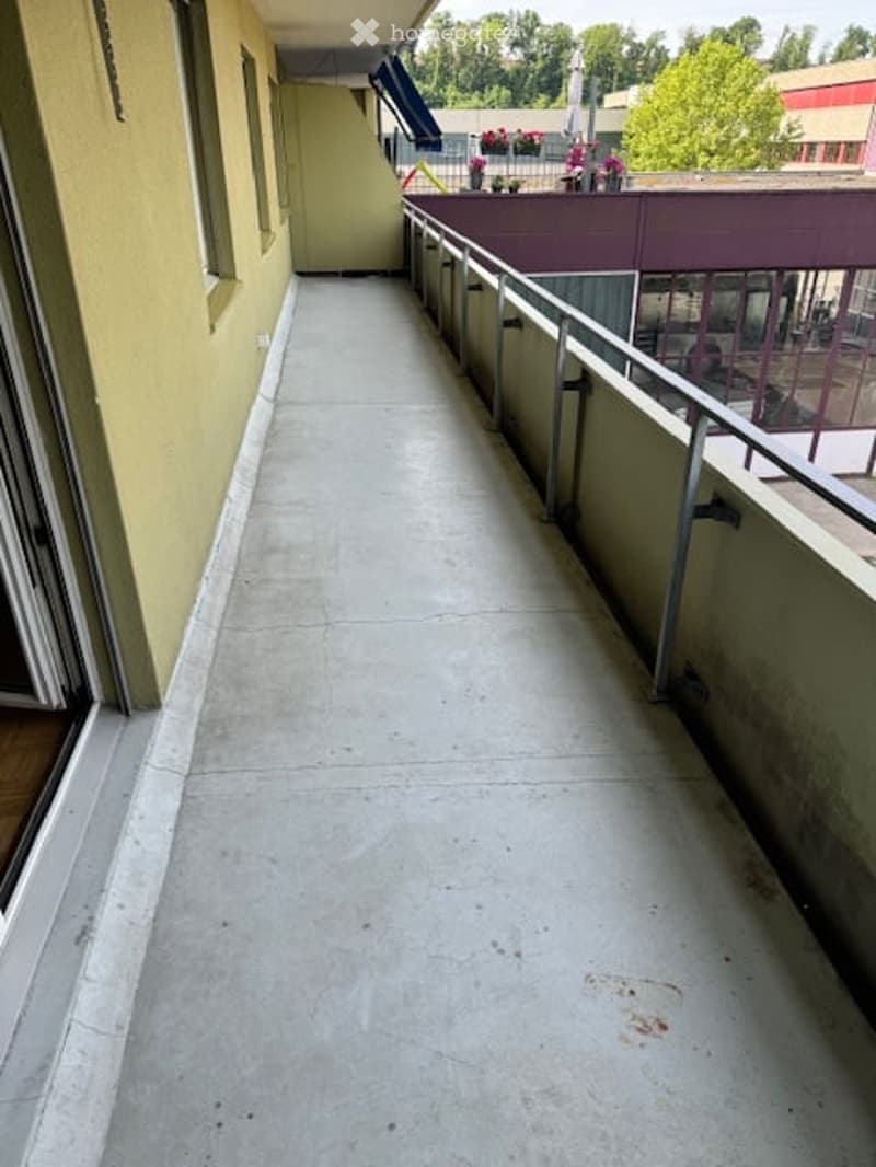 A long, narrow concrete hallway with metal railings on both sides, leading to an open area with greenery visible in the distance.