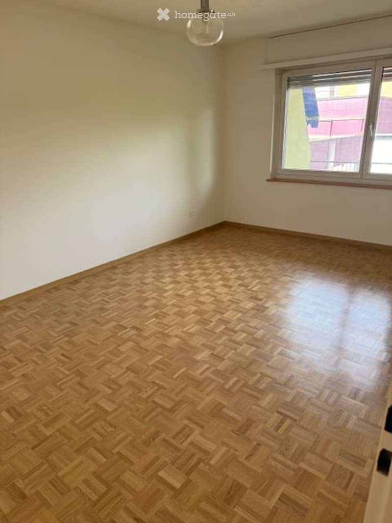 Empty room with wooden herringbone flooring, white walls, and a single window with a view of a building outside. The room has a simple, clean design with no furniture or decorations.