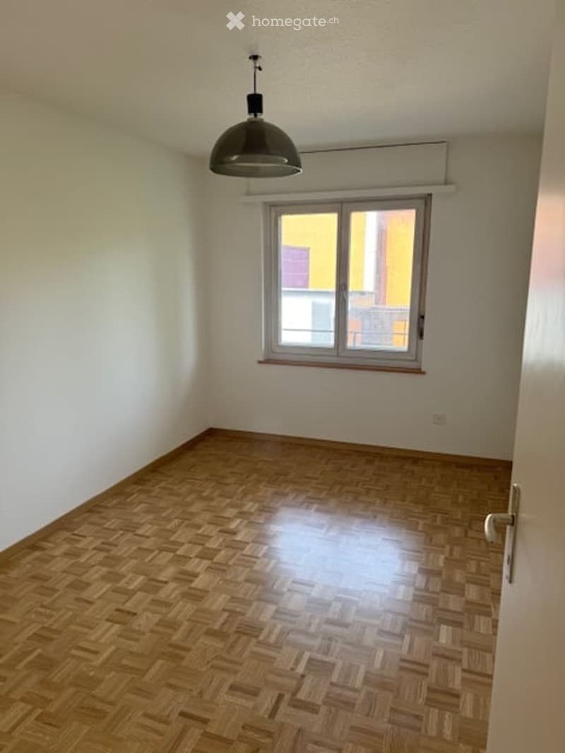 Empty room with wooden parquet flooring, white walls, and a single window with a view of a neighboring building. A pendant light fixture hangs from the ceiling.