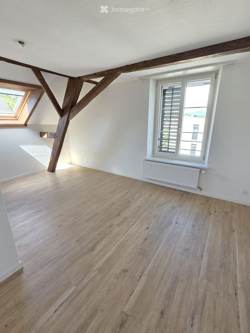 Empty room with wooden flooring, white walls, exposed wooden beams, and a window with blinds. A radiator is visible under the window.