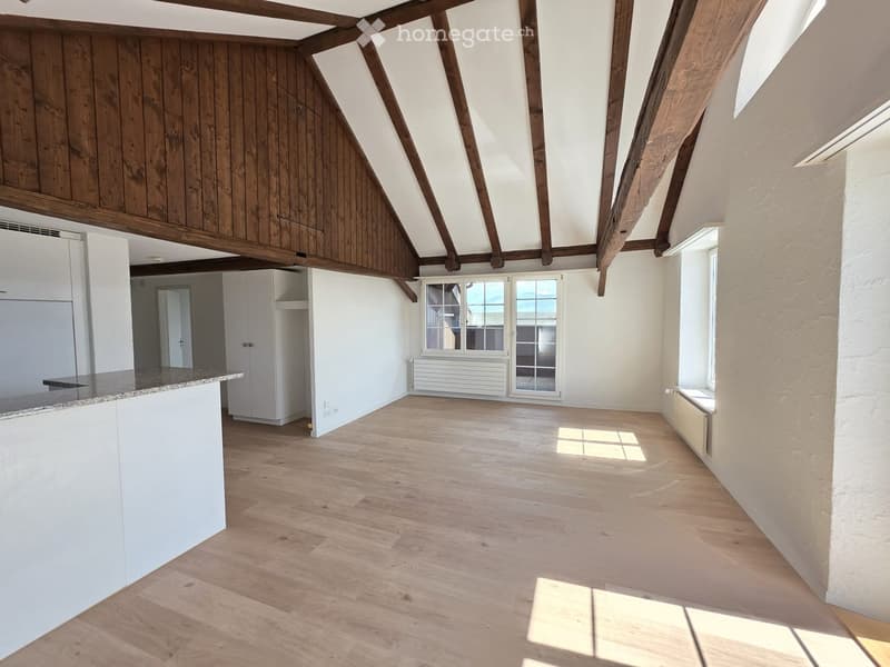 Empty attic space with wooden beams, white walls, wooden flooring, and large windows allowing natural light. Features a white cabinet and a countertop area.