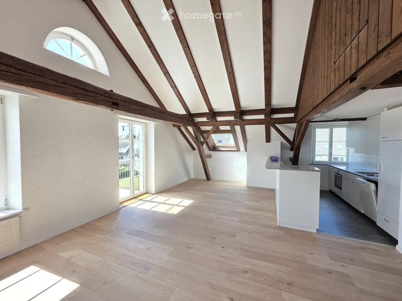 Bright, open-plan living and dining area with exposed wooden beams, light wooden flooring, and a modern white kitchen with built-in appliances.
