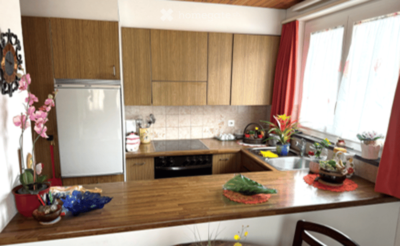 Bright kitchen with wooden cabinets, stainless steel appliances, and a central island with a wooden countertop. The space features a sink, refrigerator, and various decorative items including flower pots and a bowl on the counter.