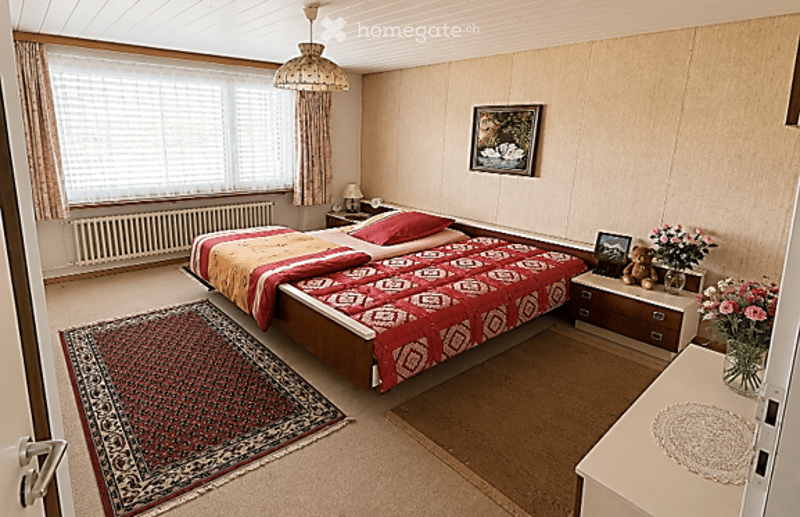 Cozy bedroom with a large bed, red and yellow bedding, nightstand with lamp, picture frame on wall, flower vase on dresser, and patterned rug on floor.