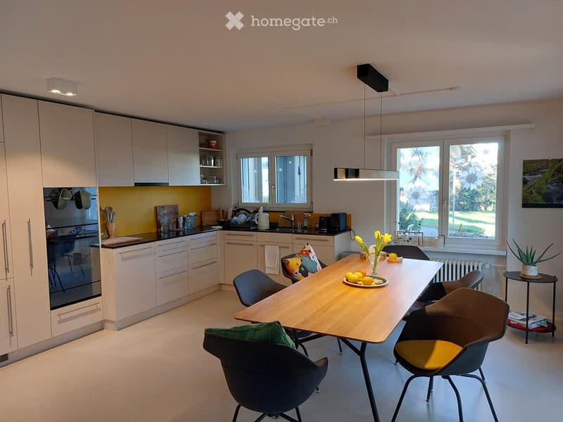 Modern kitchen with white cabinets, a central wooden dining table, four chairs, and various appliances including an oven and a refrigerator. The space features large windows and a bright, open layout.