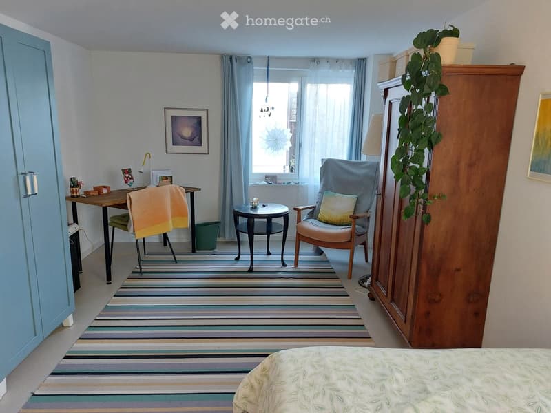 Cozy bedroom with a bed, armchair, small table, desk, and a plant. Features striped rug, large window with curtains, and a wooden cabinet.