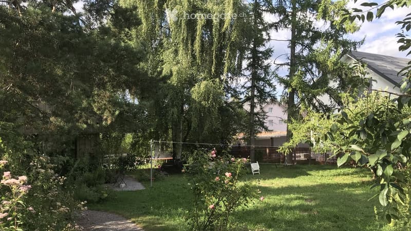 A well-maintained garden with a grassy lawn, tall trees, a gravel path, and a white folding chair. A clothesline is visible, and a fence encloses the area.